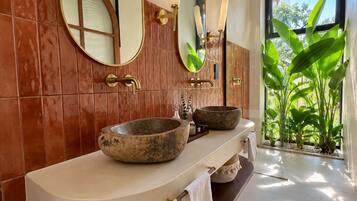 A bathroom with a large mirror, two stone basins, and a view of greenery outside.