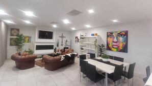 Een moderne woonkamer met een bank, eettafel en stoelen, een aan de muur bevestigde televisie en een kleurrijk schilderij.