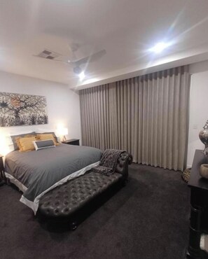 Une chambre d’hôtel comprenant un lit, un banc, un ventilateur de plafond, une grande fenêtre avec des rideaux et une œuvre d’art accrochée au mur.