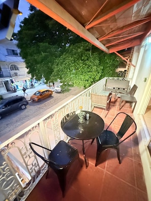 A balcony with a table and chairs, a view of a street with cars and pedestrians, and a tree in the background. - CONDO W/BALCONY 1 STREET FROM BEACH AND BOARDWALK (Puerto Vallarta)
