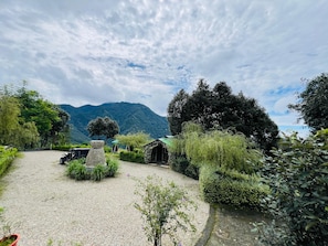 Uma trilha de cascalho leva a um edifício rústico com um pilar de pedra, cercado por vegetação exuberante e uma paisagem montanhosa.