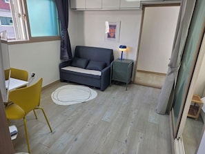 Um quarto pequeno com um sofá, uma mesa com cadeiras, um abajur e um pôster na parede.
