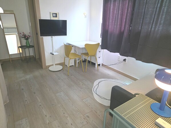 A room with a wooden floor, a desk with two yellow chairs, a TV mounted on the wall, and a lamp on a table.
