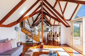 Ein großzügiger Loft mit hohen Decken, freiliegenden Holzbalken, einer Treppe, einer Wohnbereits mit einer Couch und Blick durch eine große Schiebeglastür.