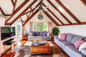 Ein geräumiges Wohnzimmer mit hoher Decke, Holzbalken, einem großen Fenster, einem Fernseher, einer Couch und einem Couchtisch.