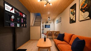 A modern living room with a TV displaying Netflix recommendations, a wooden table, an orange sofa, and a staircase leading to another room. - Rose House (Osaka Nishinari)