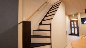 A staircase with a wooden handrail and black steps, leading to a room with a wooden floor and a door with a window. - Rose House (Osaka Nishinari)