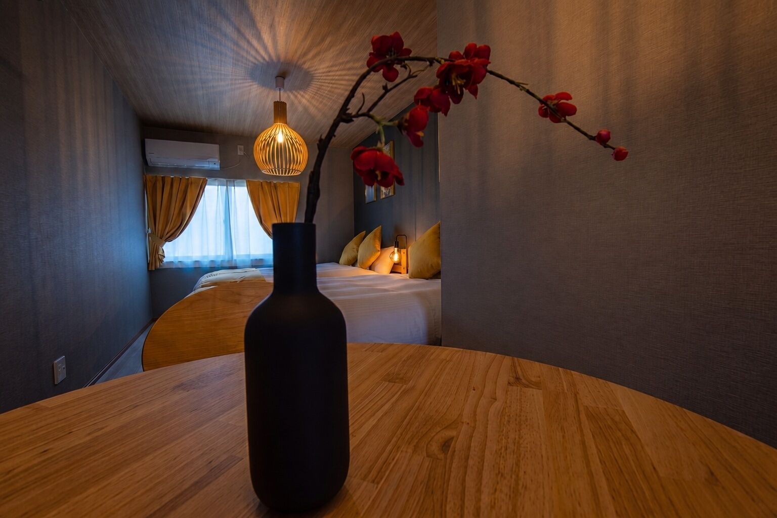 A hotel room with a bed, a bedside table with a vase, a hanging light fixture, and a view of another room.