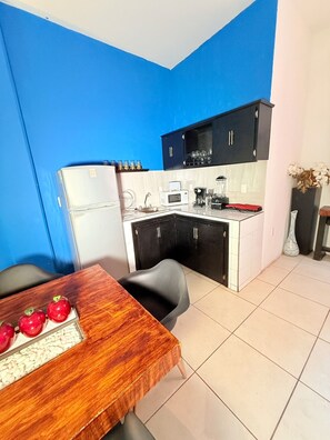 Cozinha compacta com geladeira, micro-ondas e cafeteira. Há uma mesa de jantar com uma fruteira e uma cadeira. As paredes são azuis e o chão é revestido com azulejos.