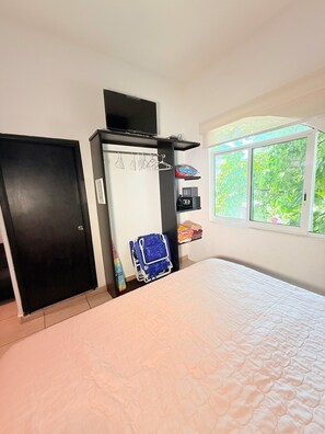 Een slaapkamer met een bed, een kledingkast, een plank met opgevouwen kleding, een blauwe stoel en een raam met uitzicht op groen.