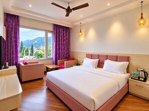 A hotel room with a large bed, a desk, a chair, a TV, and a window with a view of mountains. - Hotel Satyaki Orchid (Katra)