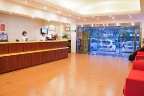 Zona de recepción de hotel con mostrador de madera, televisor y entrada de vidrio a la calle.