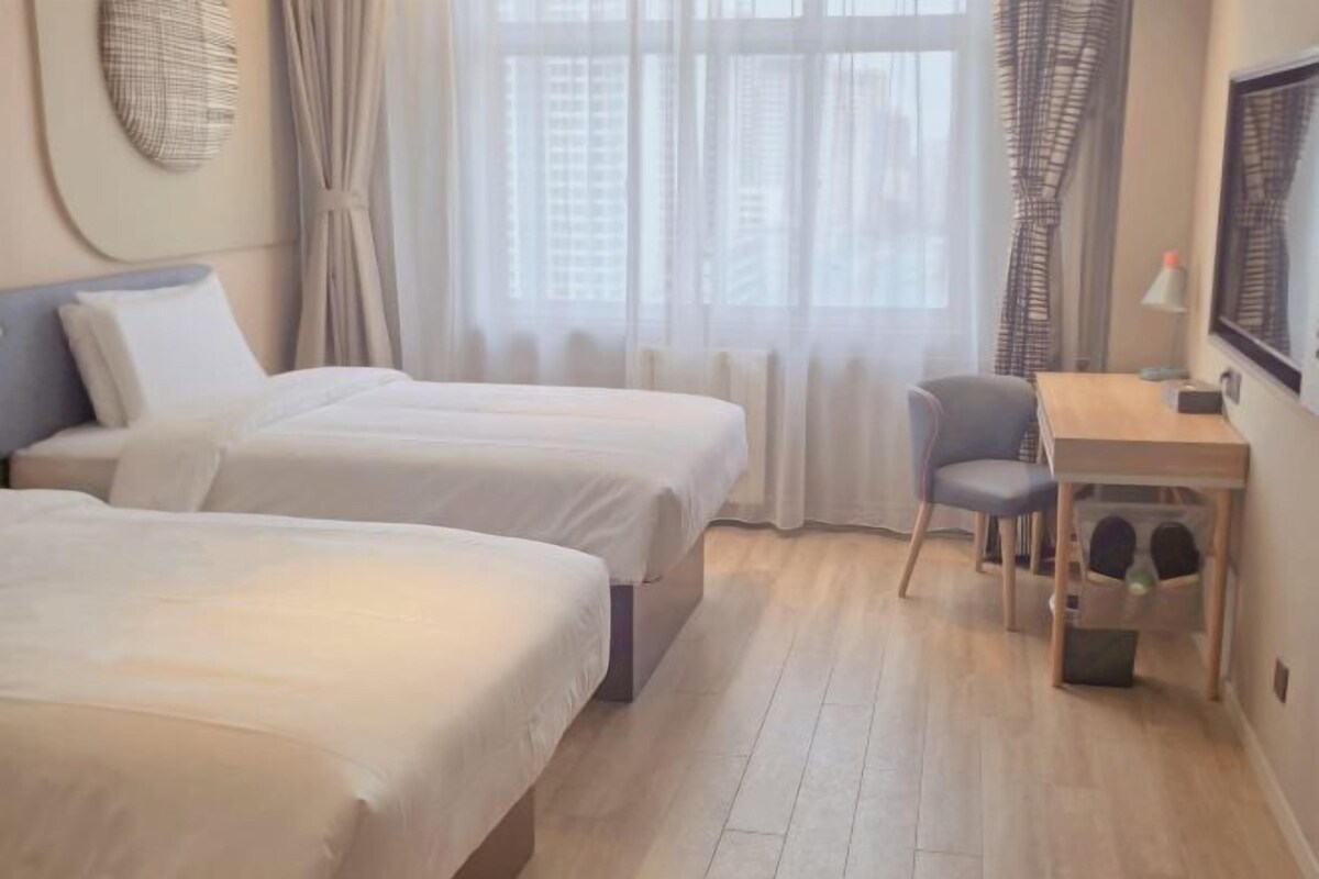 Ein Hotelzimmer mit zwei Betten, einem Schreibtisch, einem Stuhl und einem fensterbepflanzten Fenster.