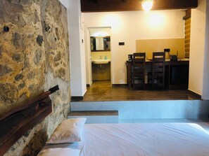 A bedroom with a stone wall, a bed with pillows, a wooden bench, and a view into a kitchen area with a sink and a telephone. - Kithulgala Cabana Resort (Kithulgala)