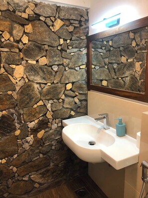 A bathroom with a stone wall, a white sink, a mirror, and a blue soap dispenser. - Kithulgala Cabana Resort (Kithulgala)