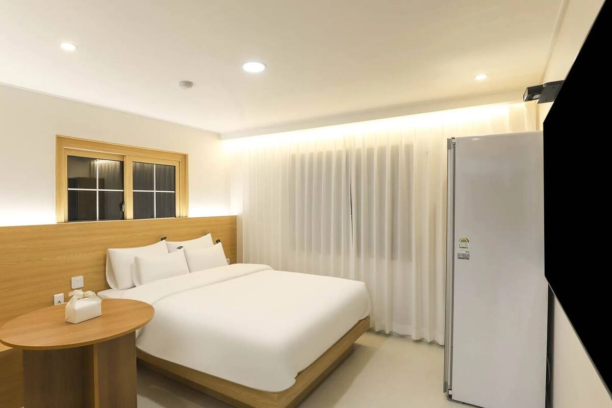 A hotel room with a bed, a small round table, a refrigerator, a window with curtains, and a wooden headboard.