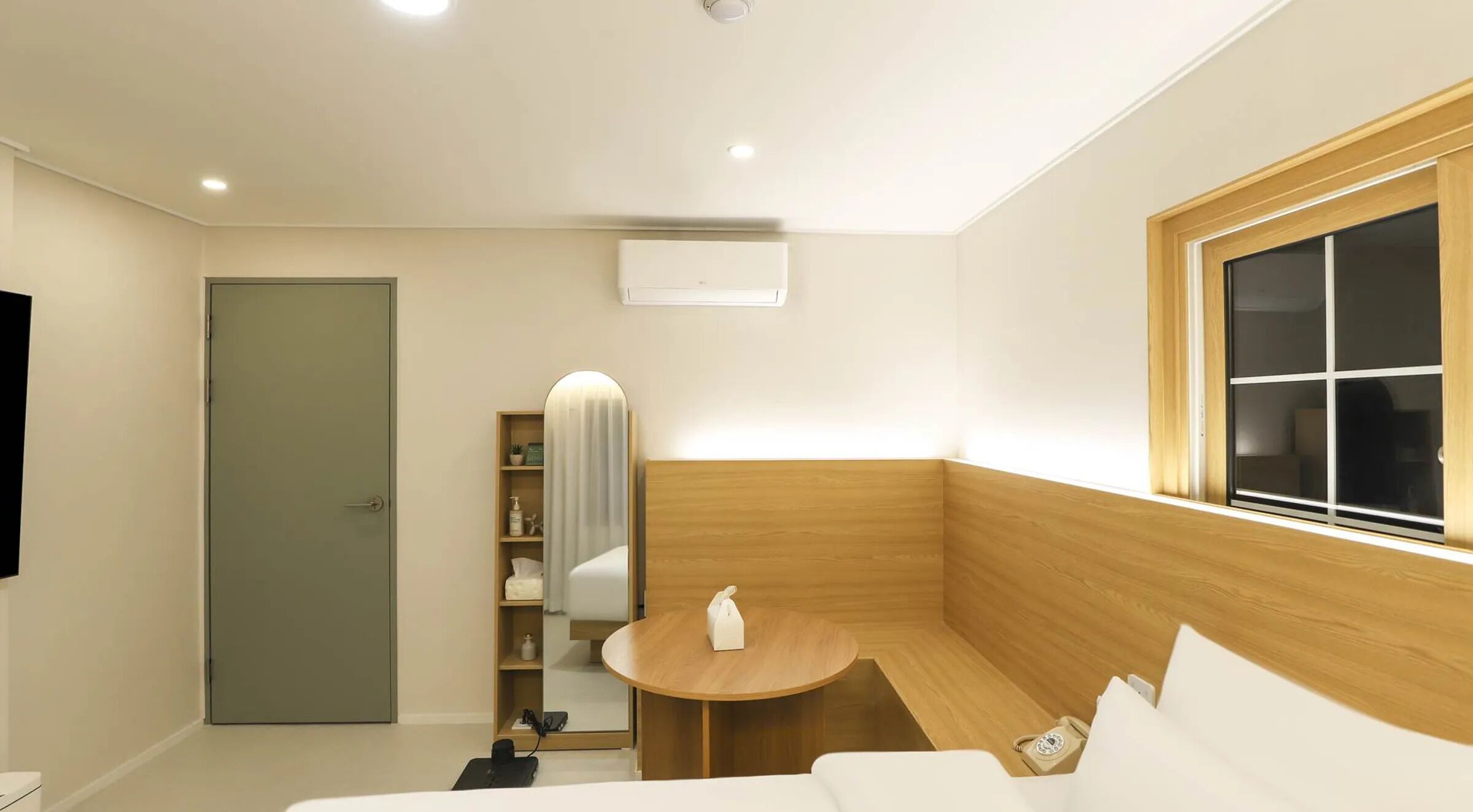 A modern hotel room with a wooden accent wall, a round wooden table, a built-in shelf with toiletries, a flat-screen TV, and a window with blinds.
