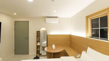 A modern hotel room with a wooden accent wall, a round wooden table, a built-in shelf with toiletries, a flat-screen TV, and a window with blinds.