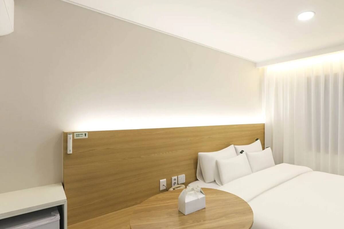A hotel room with a wooden headboard, a bed with white linens, a bedside table with a tissue box, and a window with sheer curtains.