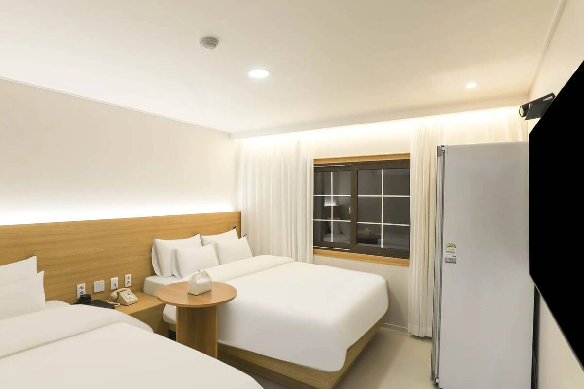 A hotel room with two beds, a bedside table with a phone, a window with curtains, and a refrigerator.