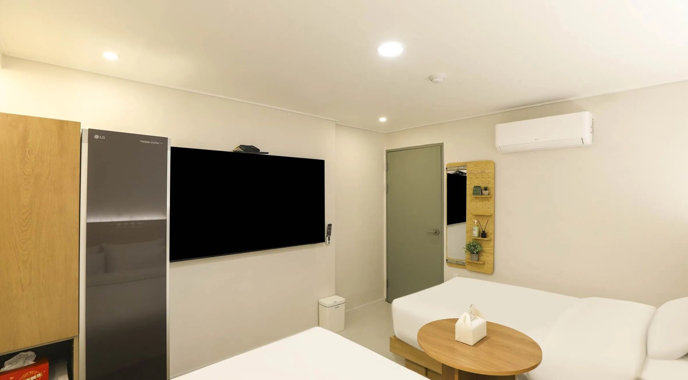 Een moderne hotelkamer met een flatscreen televisie, een houten garderobe, een klein nachtkastje, een witte bank en een spiegel aan de muur.
