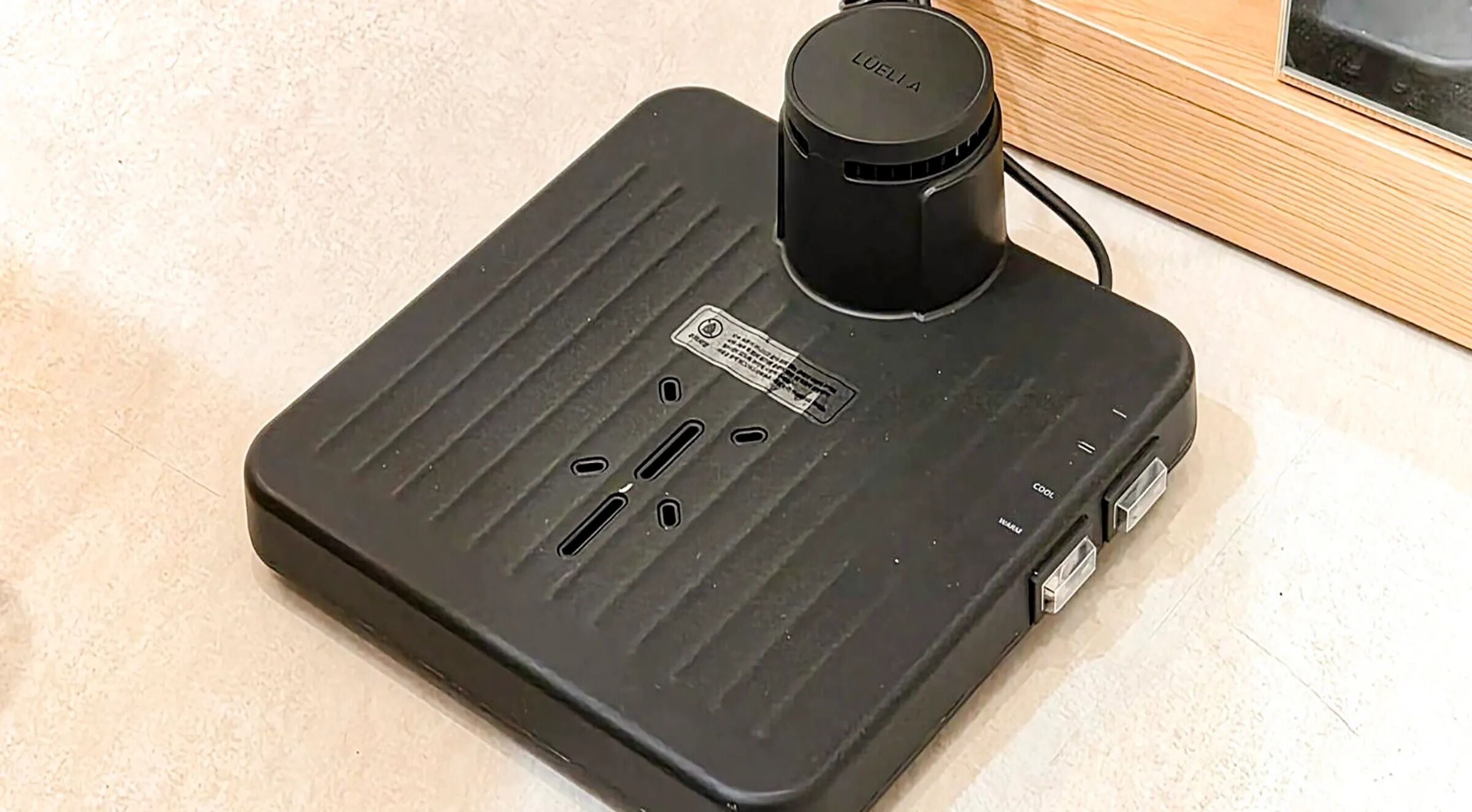 Dispositivo eletrônico preto com parte superior circular rotulada 'LUELLA', duas entradas USB e uma tomada em superfície bege.