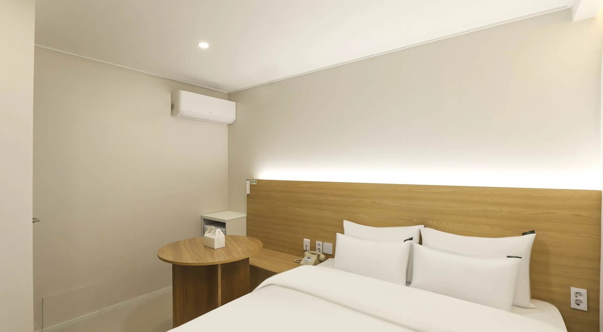 A hotel room with a bed, bedside table, nightstand, and an air conditioning unit.