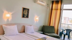 A hotel room with a bed, two pillows, a chair, a table with towels, wall-mounted lights, a painting, and a view through a window.