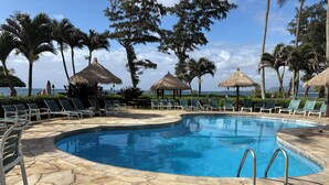 Une piscine entourée de chaises longues et de parasols en chaume, bordée de palmiers et offrant une vue sur l’océan.