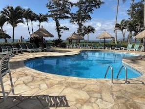 A swimming pool with lounge chairs and thatched umbrellas, surrounded by palm trees and a view of the ocean. - Tropical ocean property centrally located among beautiful floral gardens; Aloha! (Kapaʻa)