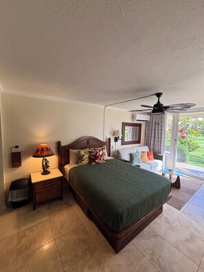 A bedroom with a bed, nightstand, lamp, ceiling fan, and a view of the outdoors. - Tropical ocean property centrally located among beautiful floral gardens; Aloha! (Kapaʻa)