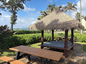 A thatched gazebo with a bench, surrounded by greenery and palm trees, overlooking a landscaped area. - Tropical ocean property centrally located among beautiful floral gardens; Aloha! (Kapaʻa)