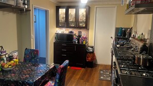 A compact kitchen with a microwave, stove, and various appliances. There is a dining table with chairs and a small trash bin. - Cheap Room in Parafield Gardens (Parafield Gardens)