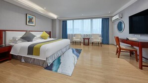 Une chambre d’hôtel comprenant un lit, deux fauteuils, un bureau, une télévision et une grande fenêtre avec des rideaux.