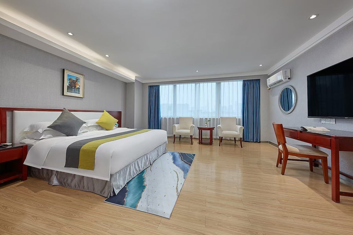 Habitación de hotel con cama, dos sillones, escritorio, televisor y ventana grande con cortinas.