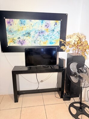 A black TV stand with a flat-screen TV, a black fan, and a decorative plant. - 2 minutes walk to the boardwalk and 4 to the beach (Puerto Vallarta)