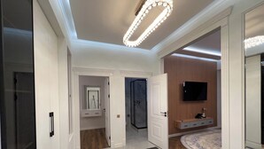 A hallway with a marble floor, a ceiling light fixture, a door leading to a bathroom, and a TV mounted on a wooden panel.