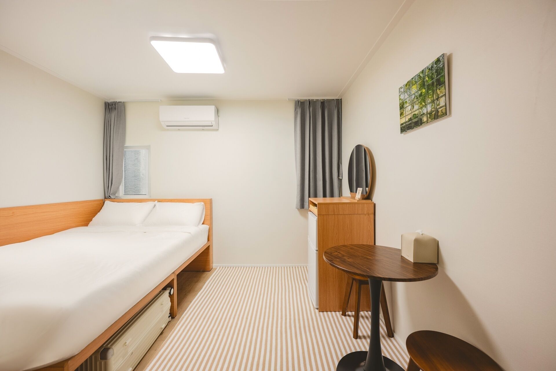 Habitación de hotel con cama, escritorio, silla, mesita, espejo, aire acondicionado y ventana con cortinas.