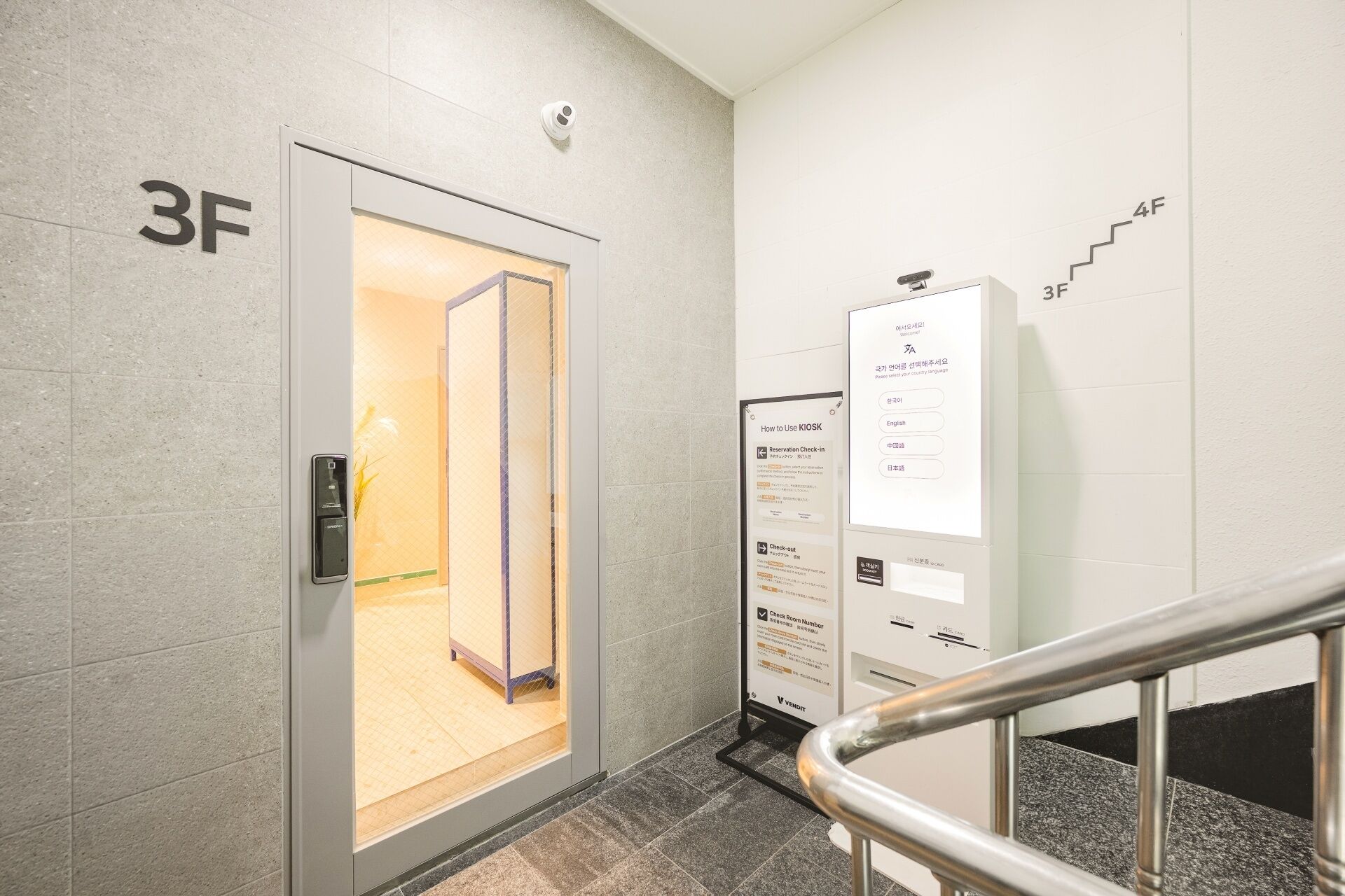 A hotel entrance with a digital kiosk for check-in, a glass door, and a staircase with a metal railing.