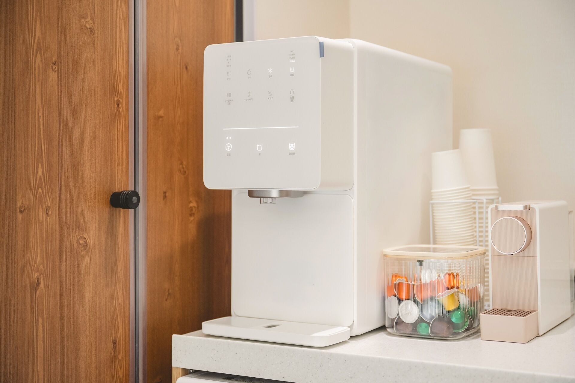 A white coffee machine with a digital display, a stack of white cups, and a glass container with colorful items on a countertop.