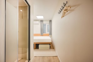 Passadís estret que condueix a un dormitori amb un llit, una finestra amb cortines i un llum penjant a la paret.