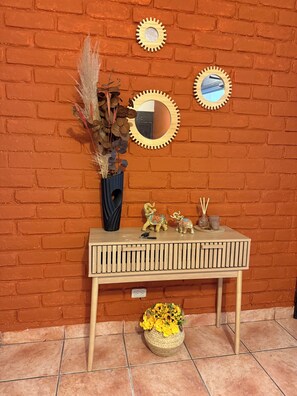 A wooden console table with decorative items, a vase with dried plants, and a woven basket of flowers against a brick wall. - Loma Linda Essenz. 152B (San Salvador)
