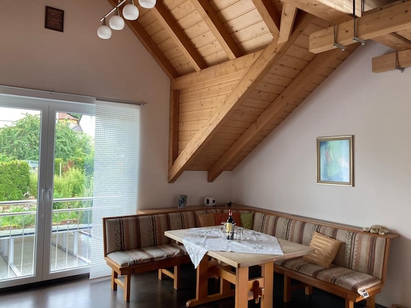 Une pièce avec un plafond en bois, une table à manger recouverte d’une nappe blanche, un banc servant de coin salon et une grande fenêtre avec des stores.