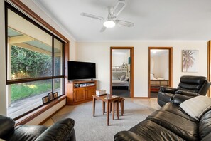 Ein Wohnzimmer mit großem Fenster, Flachbildfernseher, Ledersofa, Deckenventilator und Holzschrank.