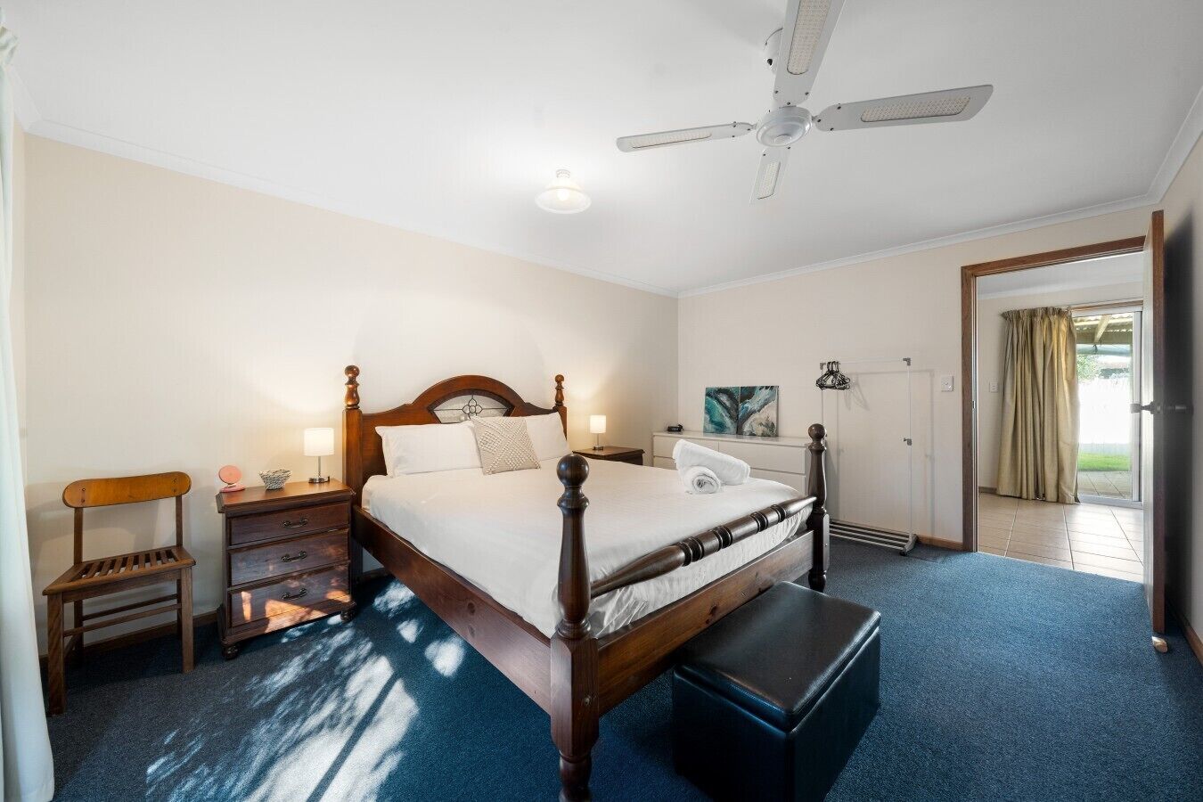 A bedroom with a wooden bed, a nightstand, a bench, a ceiling fan, and a view of the outdoors through a door.
