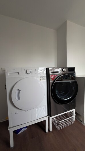 Huoneessa, jossa on puulattia ja valkoiset seinät, on kaksi päällekkäin asetettua pyykinpesukonetta.