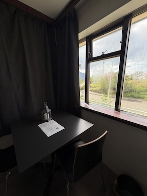 Une pièce comprenant un bureau, une chaise, une fenêtre donnant sur un espace verdoyant et un bâtiment, ainsi qu’un rideau.
