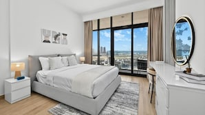 A modern bedroom with a large bed, a nightstand, a dresser, and a large window offering a city view. - Bloomfields 1BR in Azizi Riviera 3  (Dubai)