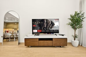 Ein modernes Wohnzimmer mit einem Flachbildfernseher, der eine Netflix-Serie zeigt, einer Holzkommode, einer Topfpflanze und einem Essbereich, der durch einen Bogen erreichbar ist.