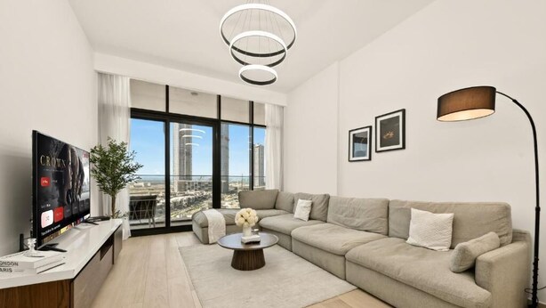 A modern living room with a large window, a sofa, a coffee table, a TV, and a lamp. - Bloomfields 1BR in Azizi Riviera 3  (Dubai)